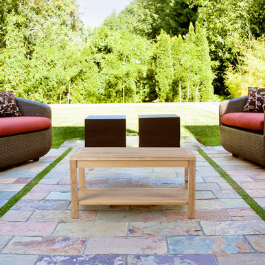 Teak outdoor patio coffee table with lower shelf on stone patio between wicker lounge chairs